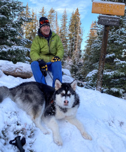 A smiling man wearing a green winter coat sitting on a fallen log on snowy ground and surrounded by snowy trees. Next to him is a sign that reads 'Homestead Trail'. In front of him is a large Husky on a leash