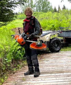 A smiling woman standing on a trail, wearing protective gear and carrying a large power tool