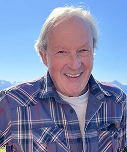 A smiling grey-haired man wearing a blue plaid shirt. Behind him is a bright blue sky.