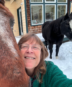 A red-haired woman with glasses, smiling. Next to her is the head of a large brown horse. Behind her is another horse, this one black with a white stripe down its face