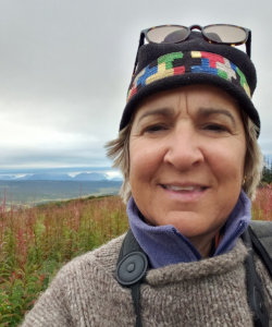 A woman in a colorful hat and a layered jacket, standing in front of a field of fireweed