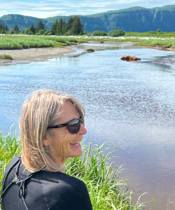 A smiling blonde woman wearing sunglasses, looking out over a river